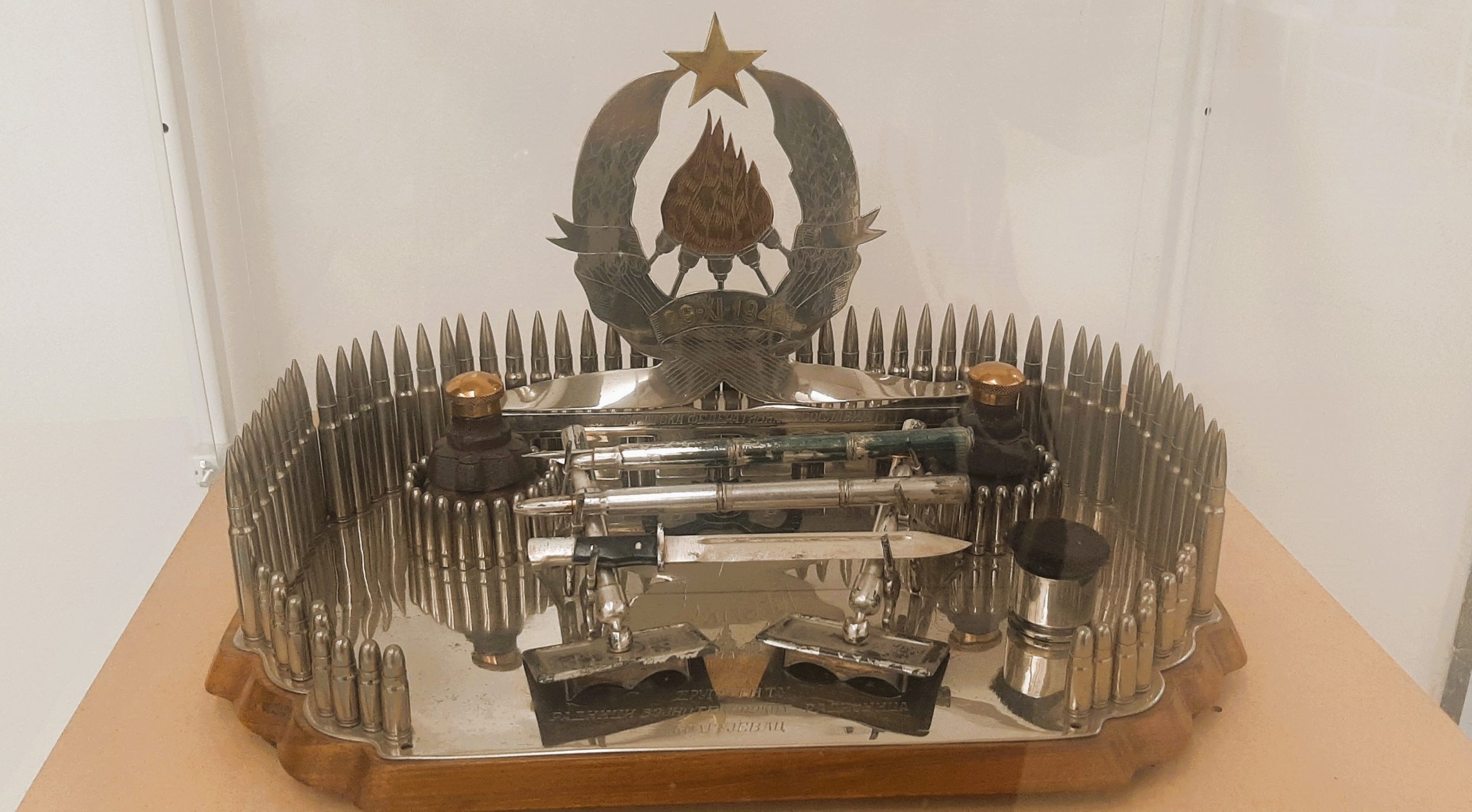 Ovo je mastionica, poklon drugu Titu za rođendan 1945 godine, koju su kreirali i napravili radnici Vojno-tehničkih radionica iz Kragujevca: meci, bočice za mastilo kao bombe, noževi (za papir!). Mastionica se nalazi u Muzeju Jugoslavije u okviru postavke izložbe poklona koje dobijao Josip Broz Tito i štafeta koje su mu uručivane za Dan mladosti. Fotografisala sam “mastionicu” 5. aprila 2025.g. u muzeju, šokirana idejom da se od metaka napravi poklon-mastionica 1945.g., iako razumem pristup kreatora da poklone ono što proizvode. Nažalost, iz današnje perspektive, SFRJ je i završila uputrebom municije i metaka, a tu su i noževi...